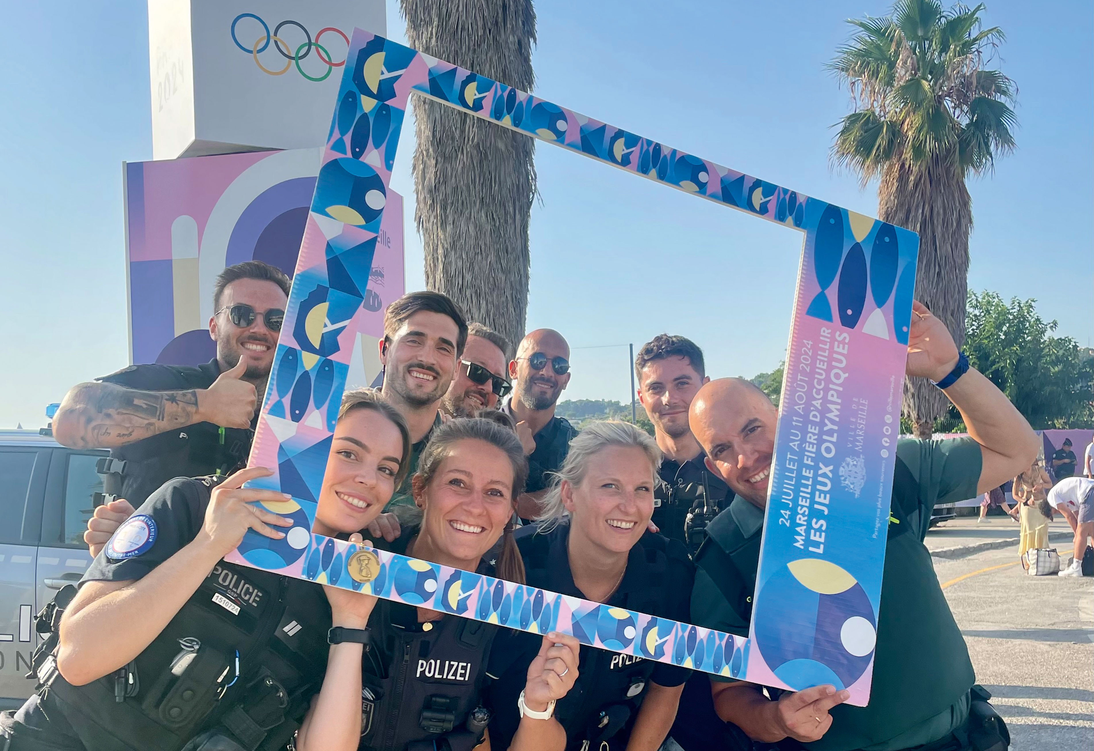 German police officer Kristina Kilb surrounded by eight French police officers at the Olympic Games in Paris. A palm tree and the Olympic rings can be seen in the background. Together they hold a photo frame through which they can see
