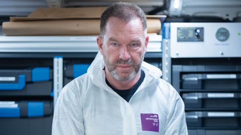 Detective Chief Inspector Frank Trojan wears a white protective suit and holds a plastic suitcase in both hands. Shelves with other suitcases and other objects can be seen in the background.